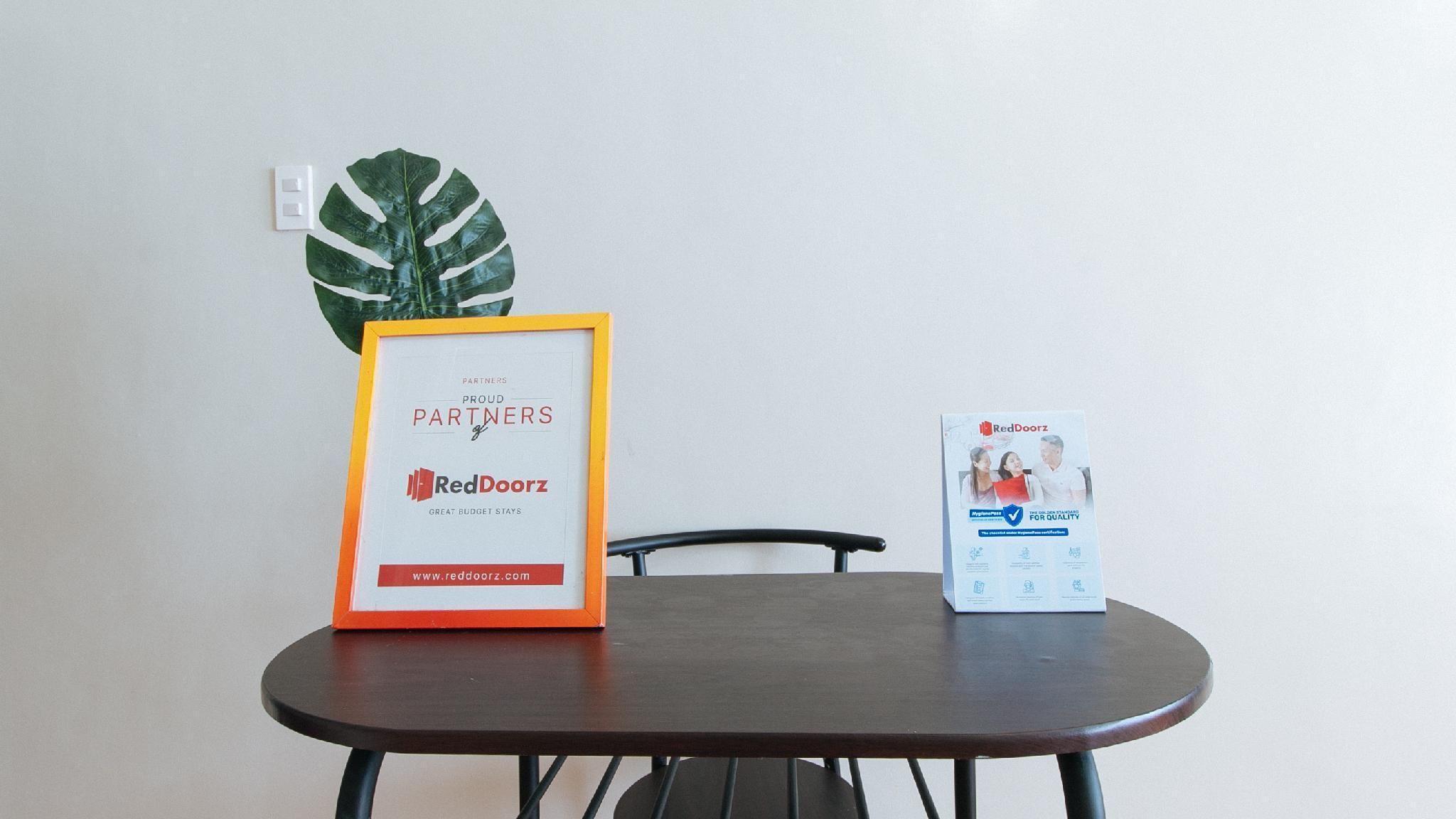 A framed sign on a table with a RedDoorz logo and a website URL, a potted plant, and a RedDoorz brochure.