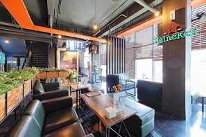 A modern restaurant interior with a Heineken sign, wooden seating, and a staircase leading to an upper level. - Sans Hotel Roxy Jakarta (Jakarta)