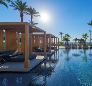 A poolside area with lounge chairs and wooden pergolas, overlooking a clear blue sky and calm water. - Ammades All Suites Beach Hotel - Adults Only (Rhodes)