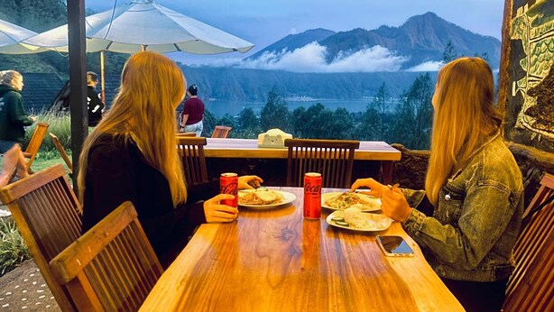 A wooden table with chairs, two people dining, a Coca-Cola can, and a scenic view of mountains and clouds. - Bunbulan Cottage Lake View (Kintamani)
