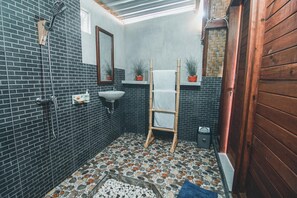 A bathroom with a tiled floor, a wooden door, a sink, a mirror, and a shower.
