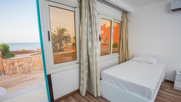 A bedroom with a bed, a window with curtains, a view of the sea, and a stone wall outside. - Lilly Rooms And Studios (Hurghada)