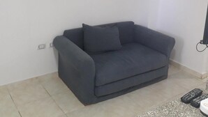 A room with a grey sofa, a painting of a desert landscape, and a TV stand with remote controls. - Lilly Rooms And Studios (Hurghada)