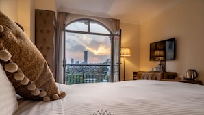 A hotel room with a bed, a lamp, a TV, and a view of the cityscape through a large window. - Suites At The Grand Palace Hotel (Addis Ababa)
