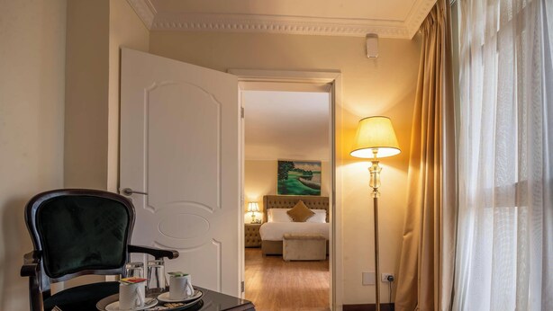 A hotel room with a bed, a bedside lamp, a chair, and a table with a cup and saucer. - Suites At The Grand Palace Hotel (Addis Ababa)