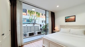 Habitación de hotel con un gran ventanal corredero, cama con ropa blanca, mesita de noche pequeña con una lámpara y un cuadro en la pared.
