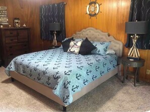 Un dormitorio con una cama que presenta un diseño de ancla náutica, cabecera de madera, decoración de timón de barco y un aparador con lámpara.