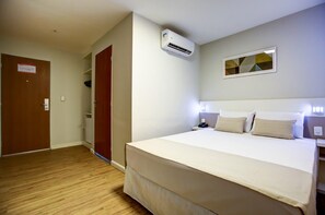 A hotel room with a bed, bedside lamps, an air conditioner, a door, and a wall-mounted light fixture.