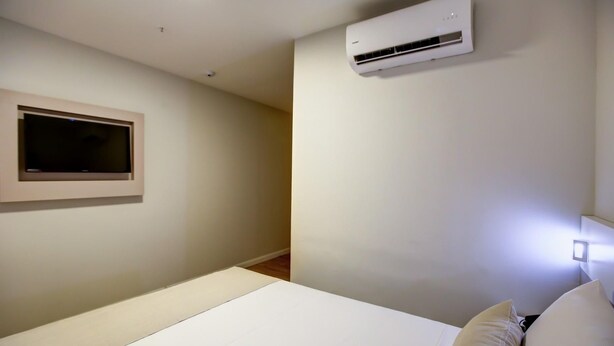 A bedroom with a bed, a mounted air conditioner, a wall-mounted TV, and a bedside lamp.