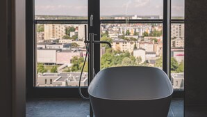 Ein modernes Badezimmer mit freistehender Badewanne, einer sichtbaren Duschbrause und Stadtblick durch große Fenster.