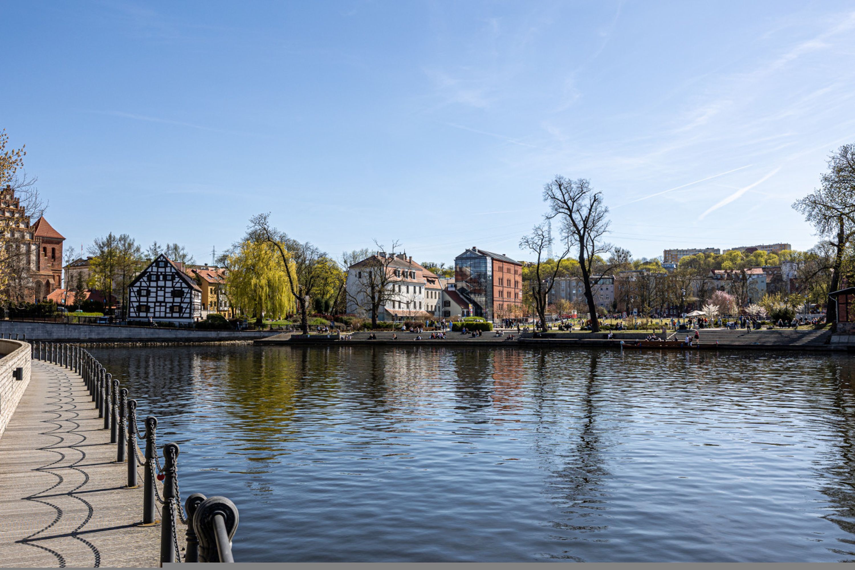A riverside walkway with buildings, a bridge, and a clear sky.