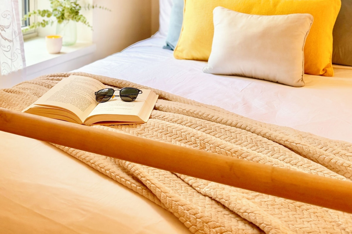 A neatly made bed with a headboard, a bedside table with a book and glasses, and a window with a plant and a candle.