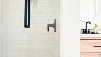 Un baño moderno con cabina de ducha de vidrio, paredes cubiertas de azulejos blancos, tocador de madera con espejo y grifo negro.