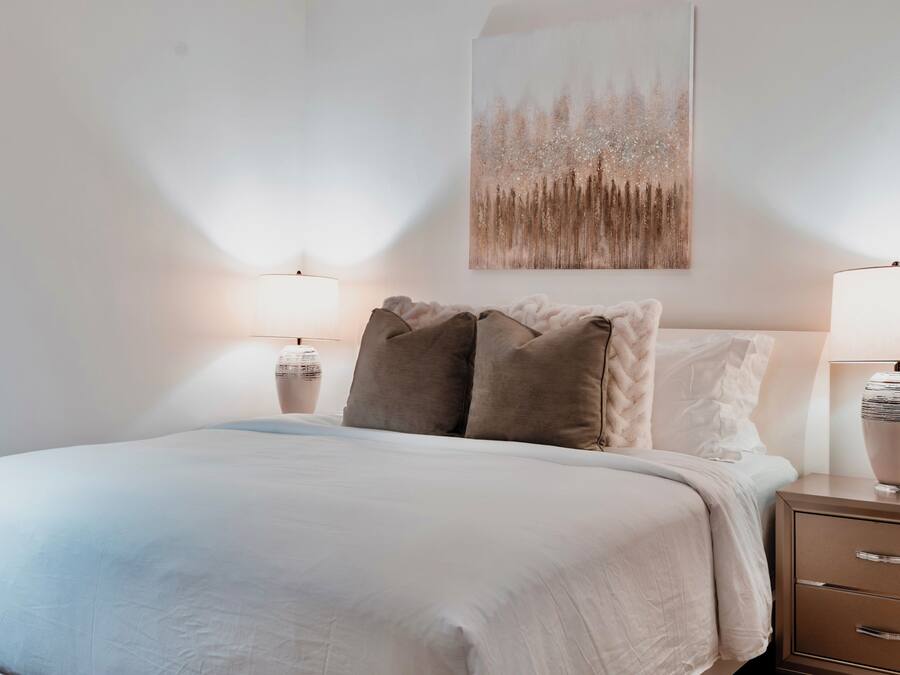 Un lit bien fait, recouvert d’une couette blanche et agrémenté de deux oreillers, avec deux tables de chevet de chaque côté, sur lesquelles sont posées des lampes.