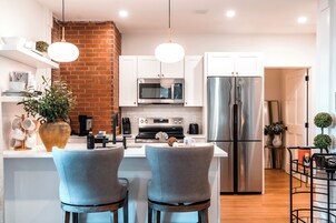Una cocina moderna con armarios blancos, un refrigerador de acero inoxidable, un microondas y una pared de ladrillo. Hay dos taburetes grises y una lámpara colgante.