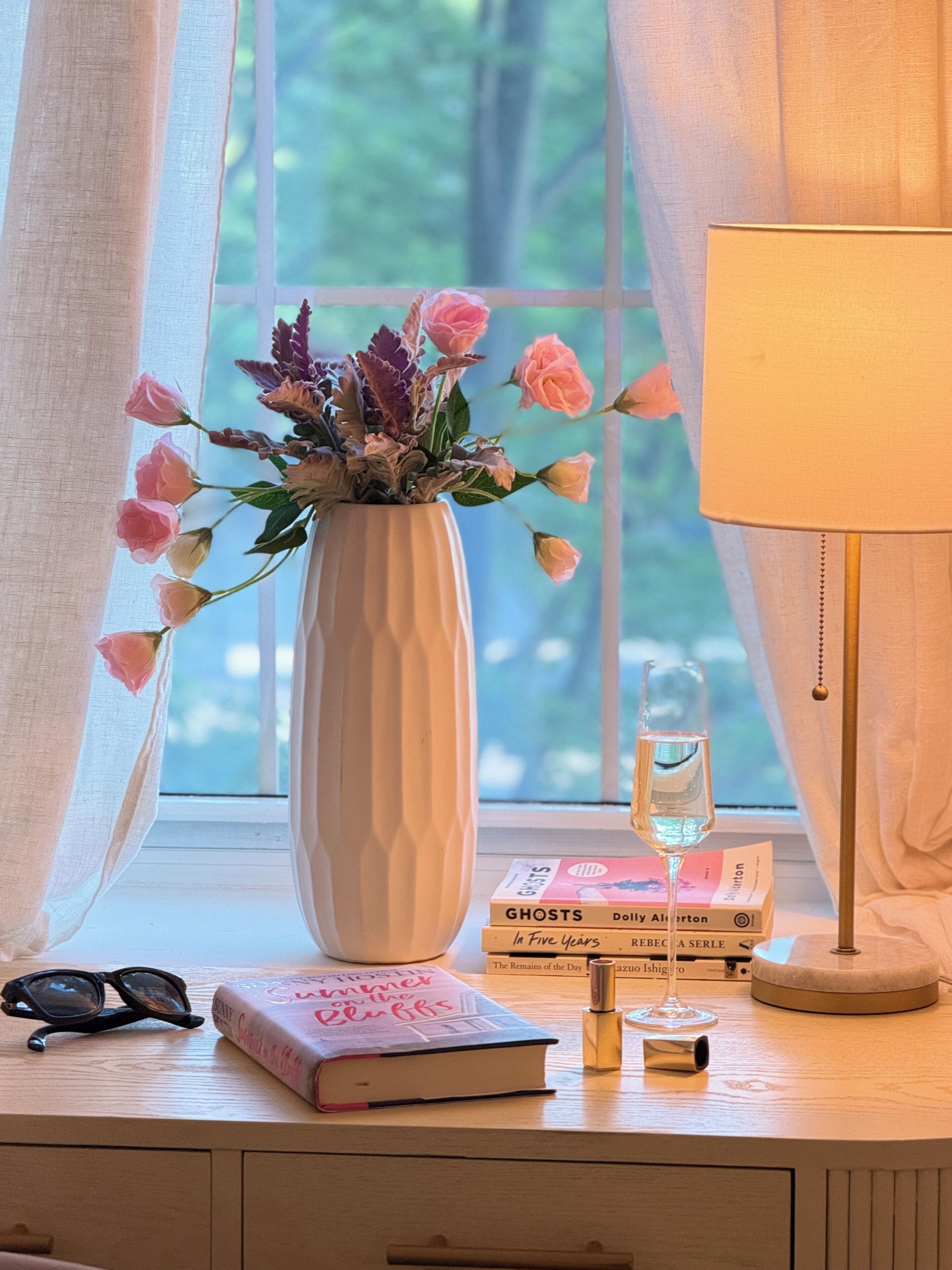 Una ventana con vistas a árboles, un jarr ón blanco con flores, una lámpara, libros y un par de gafas sobre una superficie de madera.