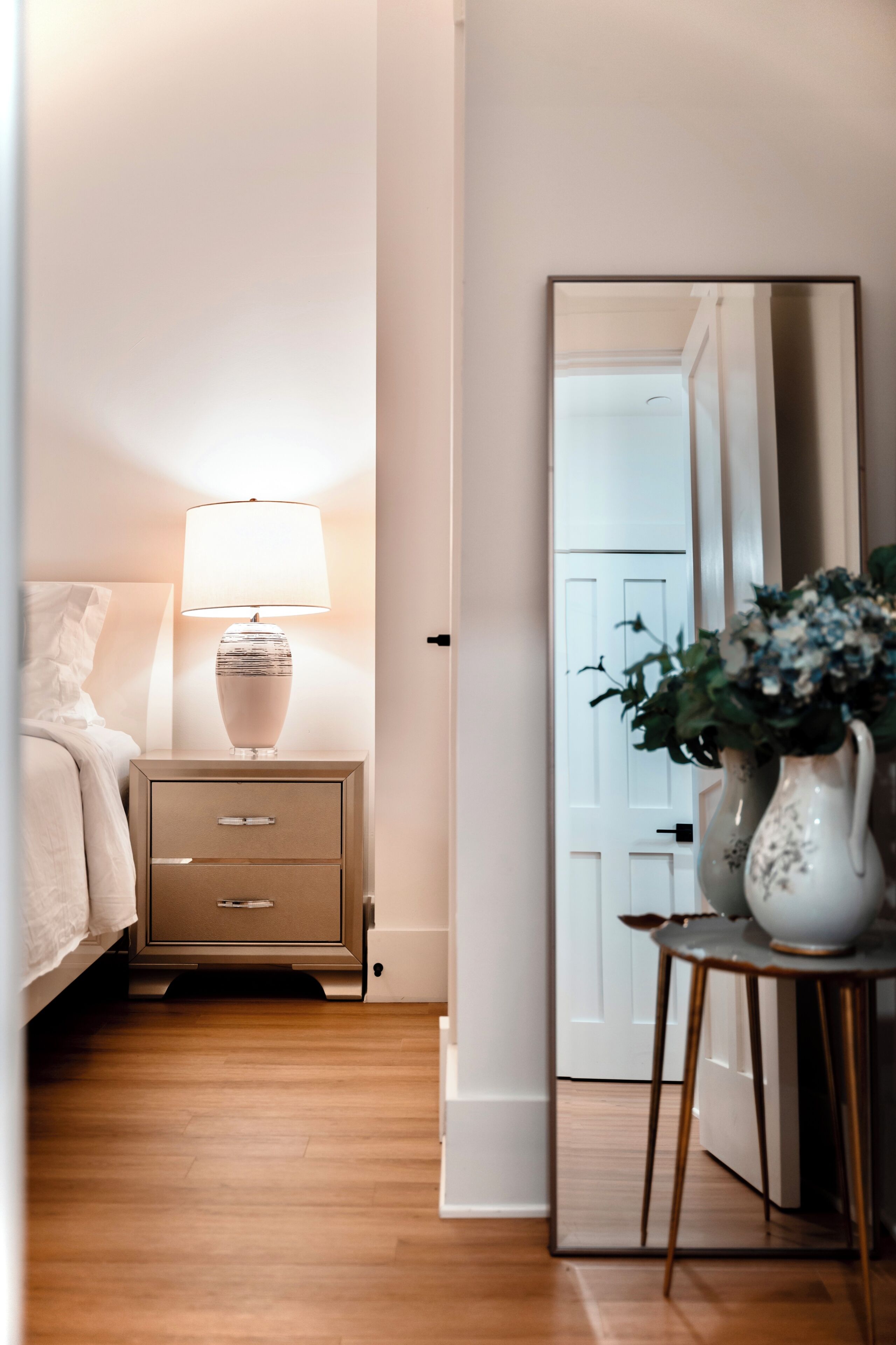 Een slaapkamer met een bed, nachtkastje met lamp, spiegel en een bloevas.