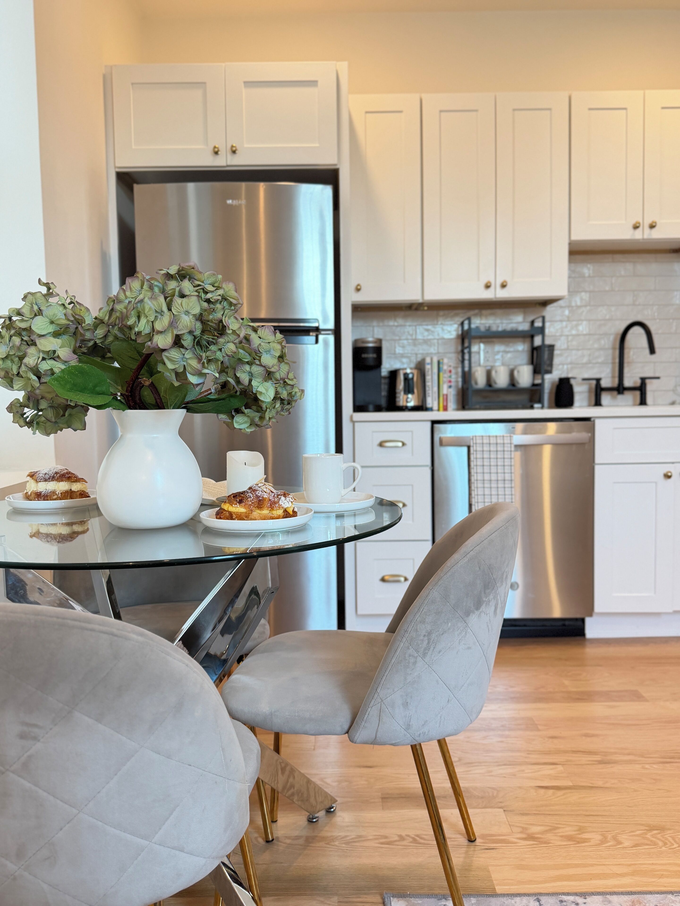 Una cocina moderna con armarios blancos, electrodomésticos de acero inoxidable, una mesa de vidrio y sillas tapizadas en gris.