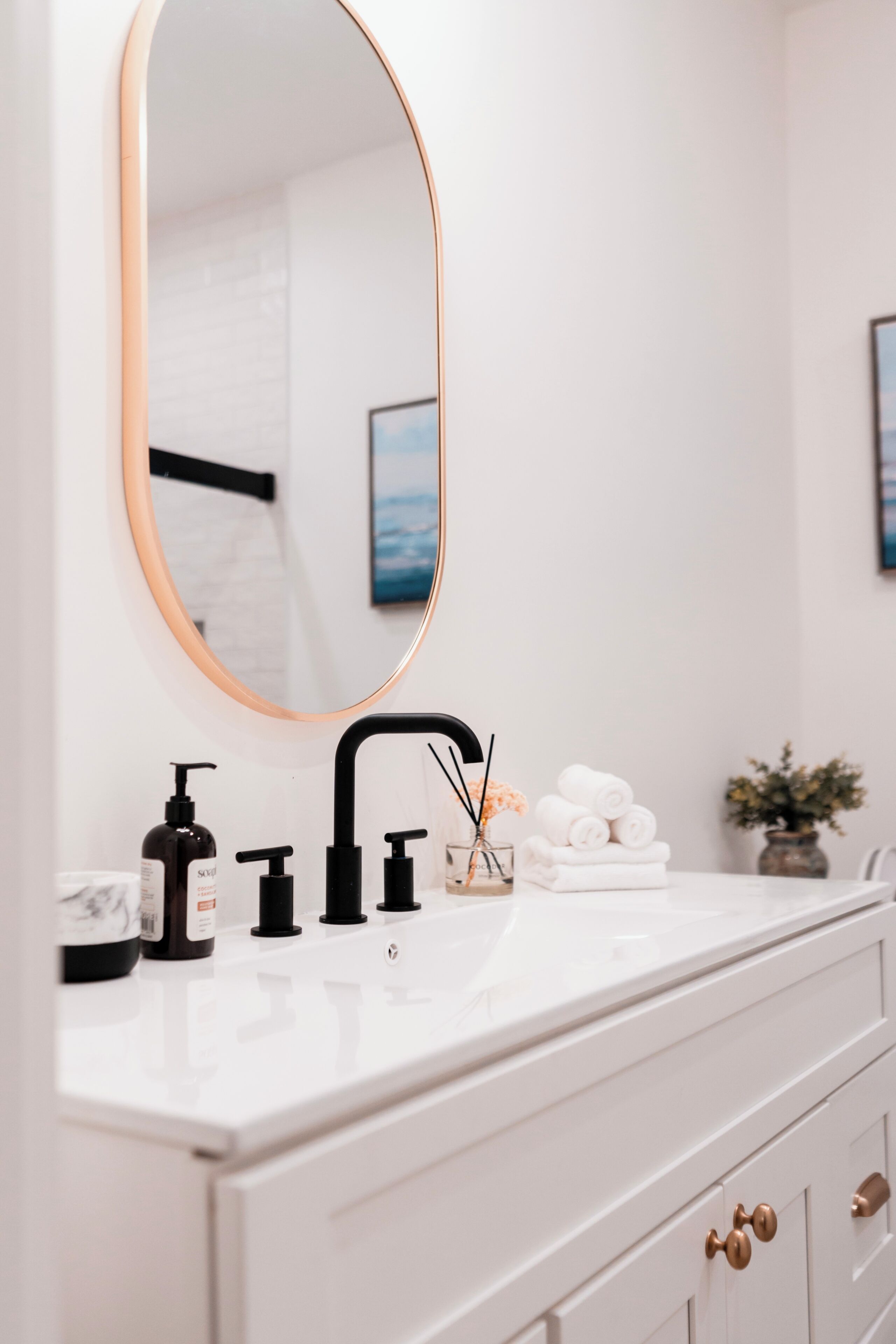 Un bagno con un mobile lavabo bianco, un rubinetto nero e uno specchio rotondo con cornice dorata. Sul lavabo ci sono articoli da toilette e un vaso con fiori.