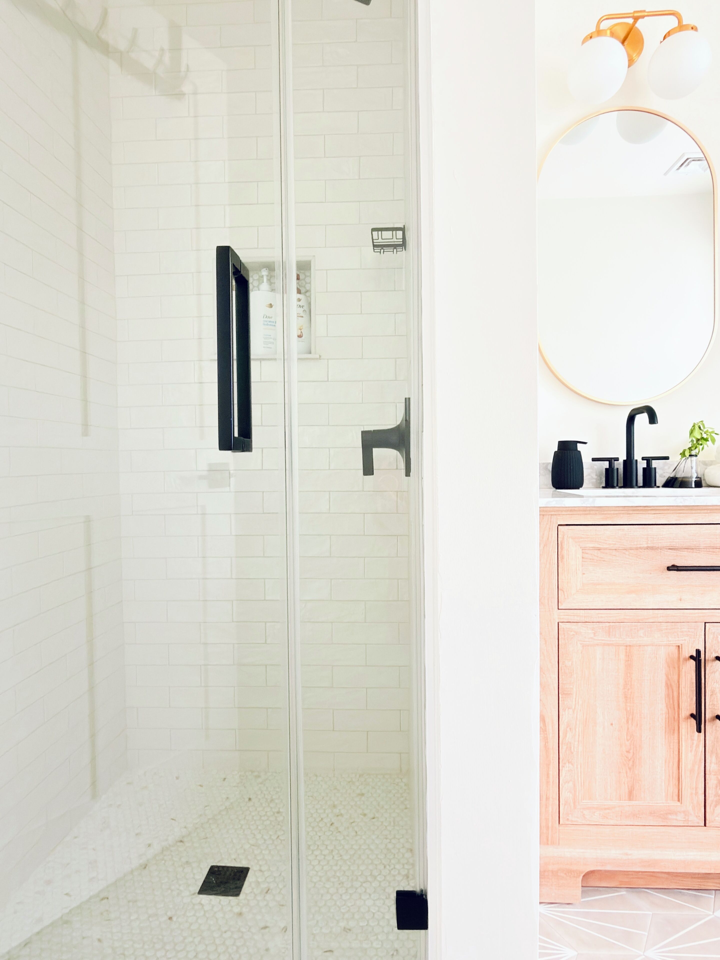 Un baño con cabina de ducha de vidrio, pared de azulejos blancos, tocador de madera con espejo y grifo negro.