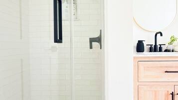 Un baño con cabina de ducha de vidrio, pared de azulejos blancos, tocador de madera con espejo y grifo negro.
