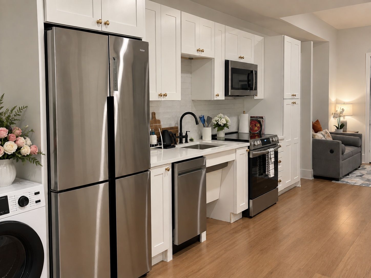A modern kitchen with stainless steel appliances, white cabinetry, and a grey sofa in the living area.