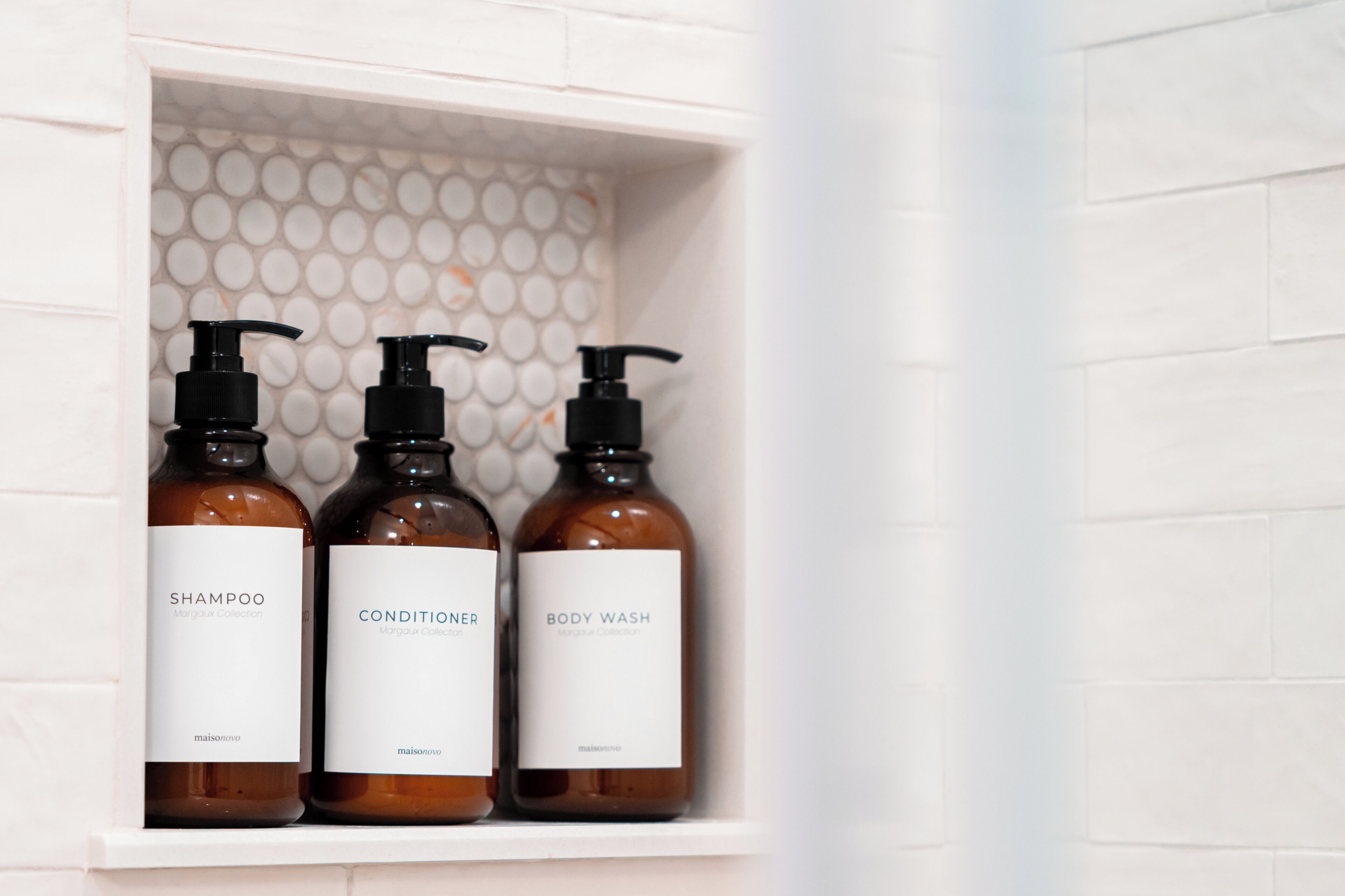A bathroom shelf with three bottles of shampoo, conditioner, and body wash from the Margaux Collection.