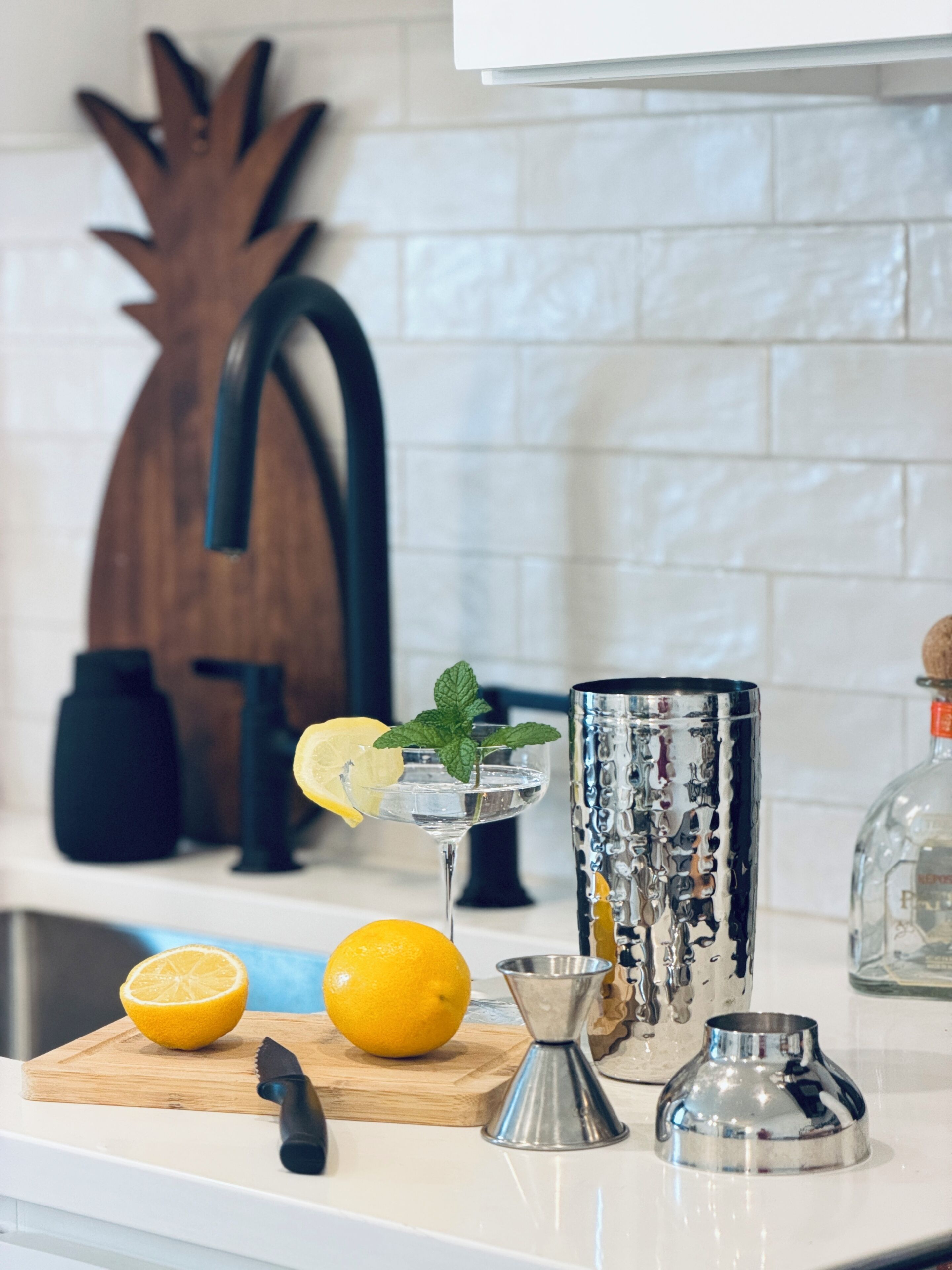 A kitchen counter with a cutting board, lemons, a knife, a cocktail shaker, and a decorative metal piece.