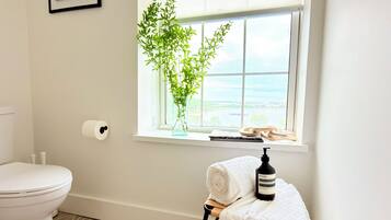Un baño con inodoro, ventana con vistas, un cuadro enmarcado y una planta.