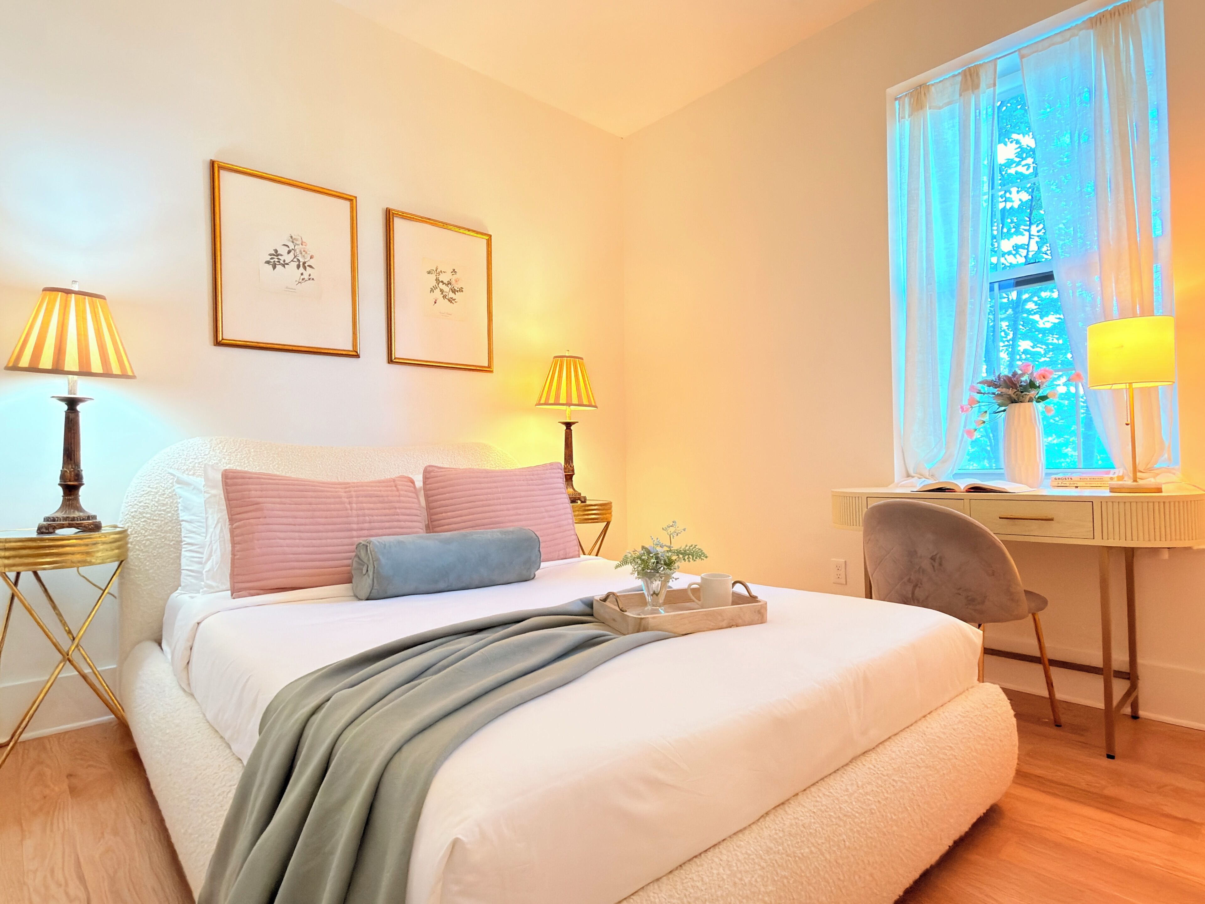 A bedroom with a bed, bedside lamps, a desk, a chair, a window with curtains, and framed pictures on the wall.