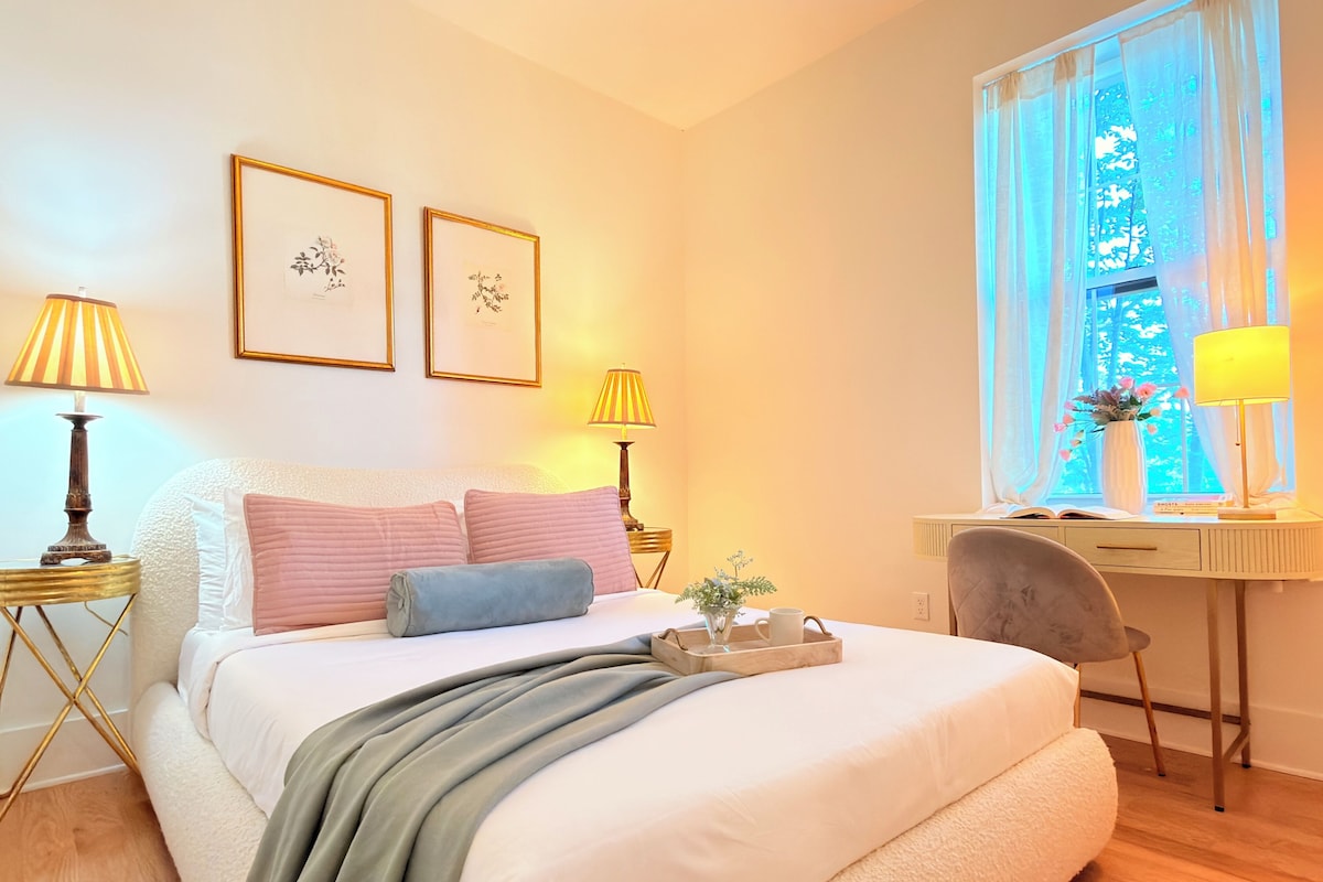 A bedroom with a bed, bedside lamps, a desk, a chair, a window with curtains, and framed pictures on the wall.