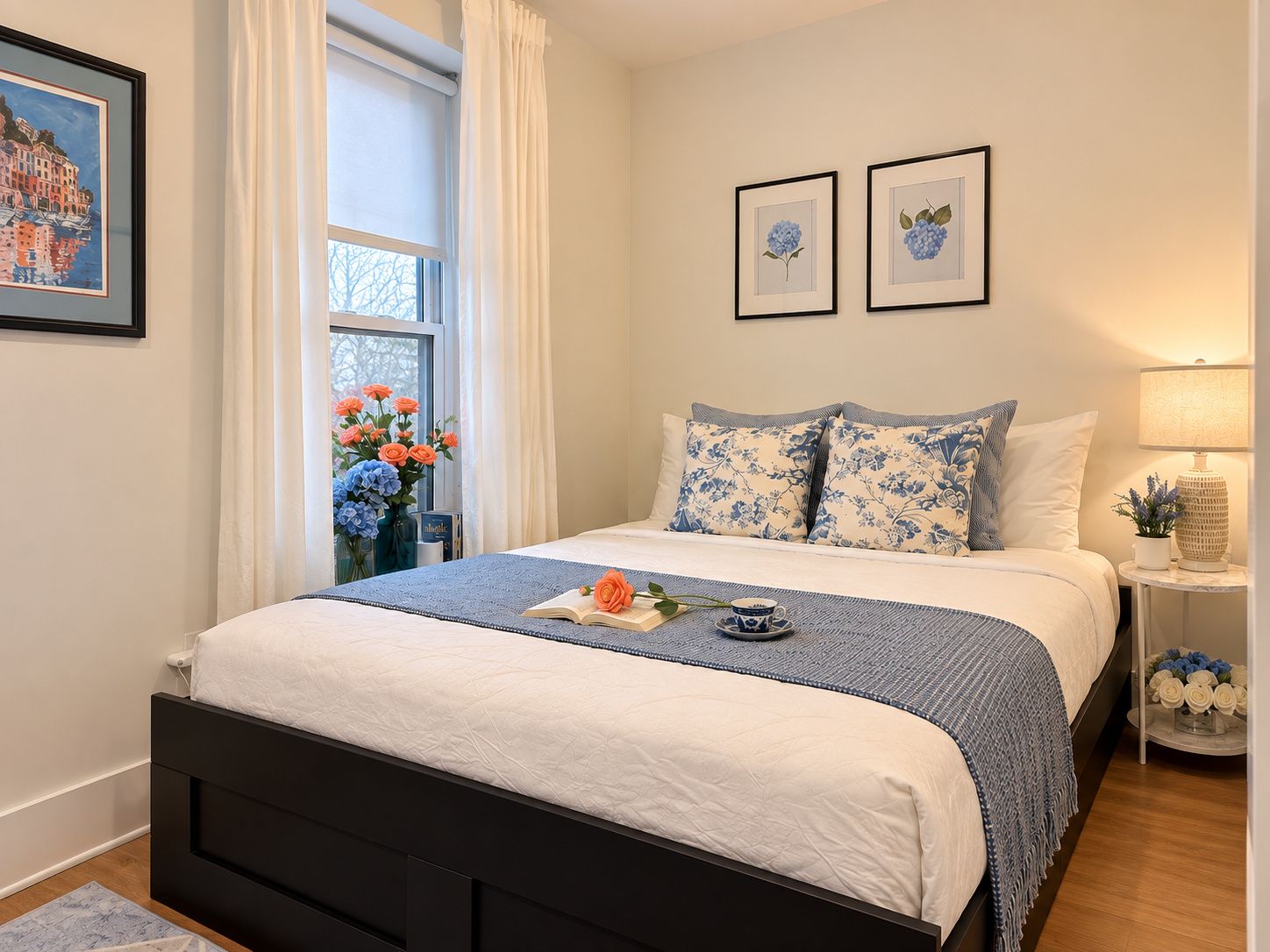 Un dormitorio ordenado con cama, ventana con cortinas, mesita de noche con lámpara y dos cuadros en la pared.