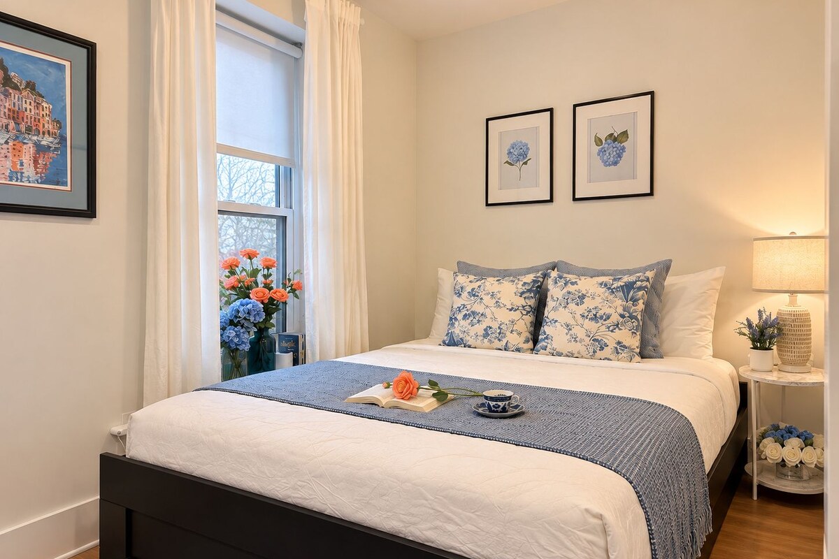 Quarto bem arrumado com cama, janela com cortinas, mesa de cabeceira com abajur e duas imagens emolduradas na parede.