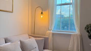 Et soveværelse med seng, puder, en sengebordlampe, et vindue med gardiner og et billede i ramme på væggen.