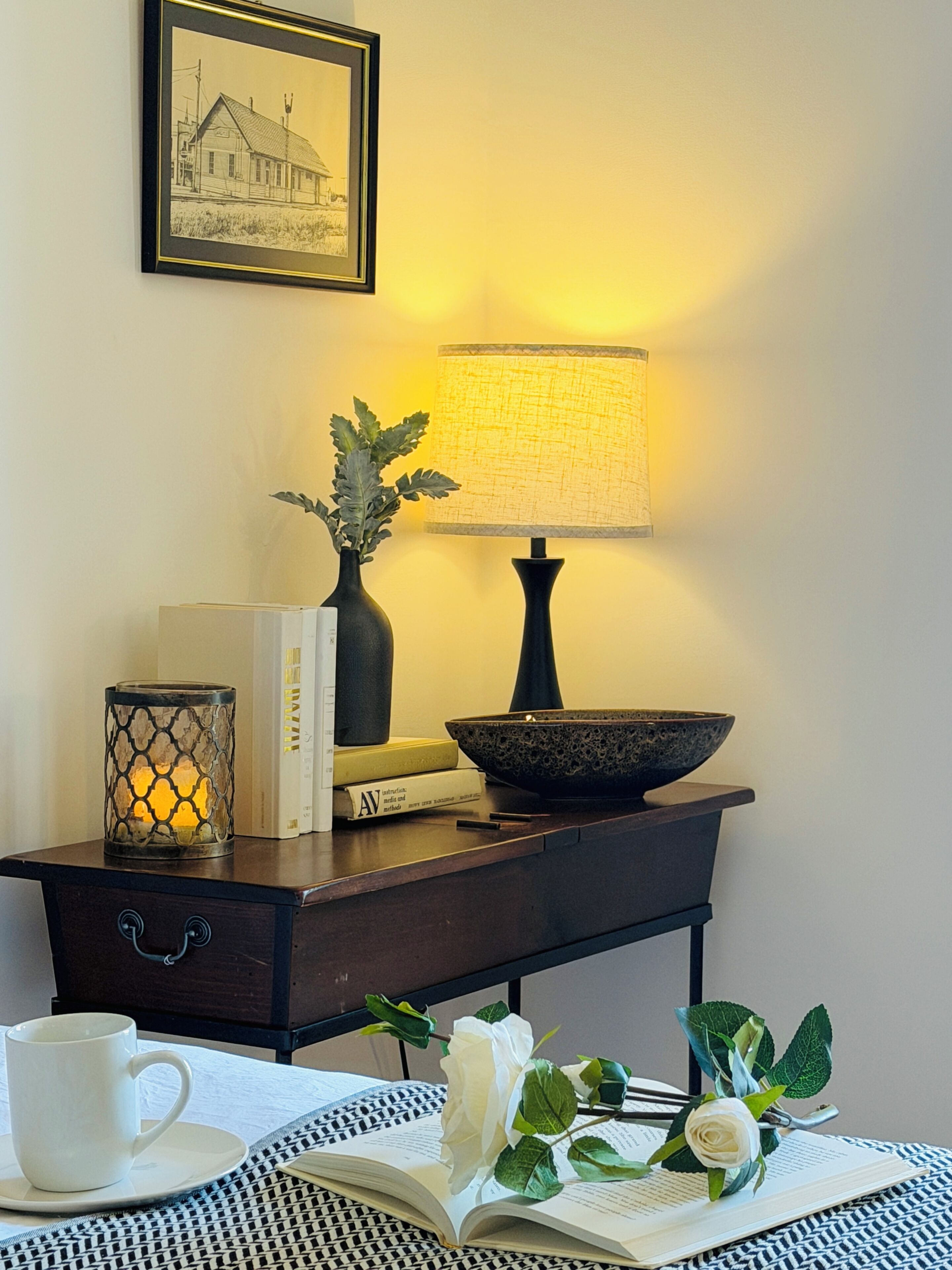 A room with a dark wooden side table, a lamp, a framed picture, books, a candle, a bowl, a cup, a rose, and an open book.