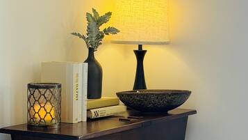 A room with a dark wooden side table, a lamp, a framed picture, books, a candle, a bowl, a cup, a rose, and an open book.