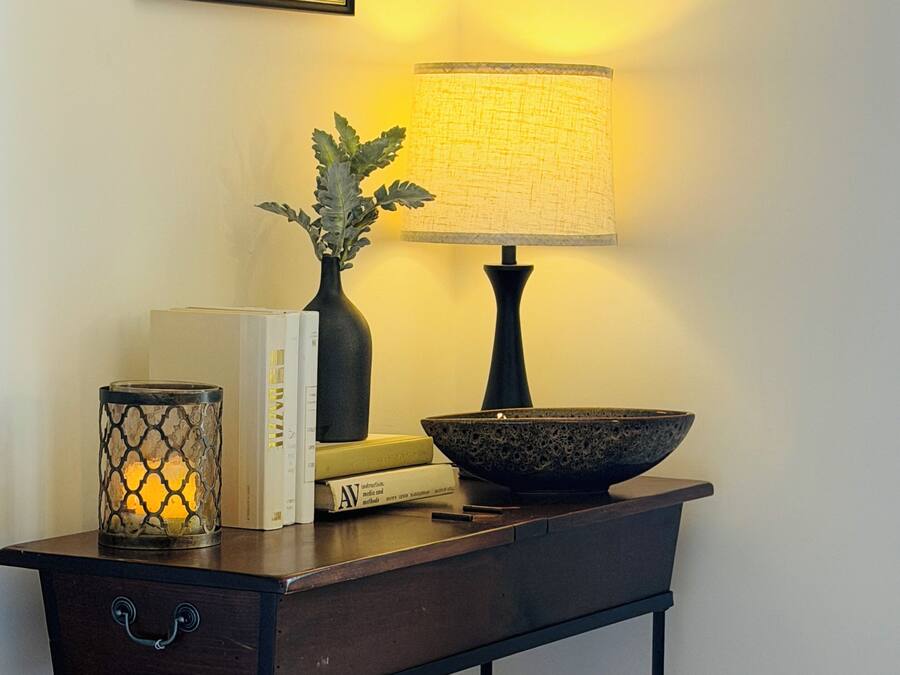 A room with a dark wooden side table, a lamp, a framed picture, books, a candle, a bowl, a cup, a rose, and an open book.