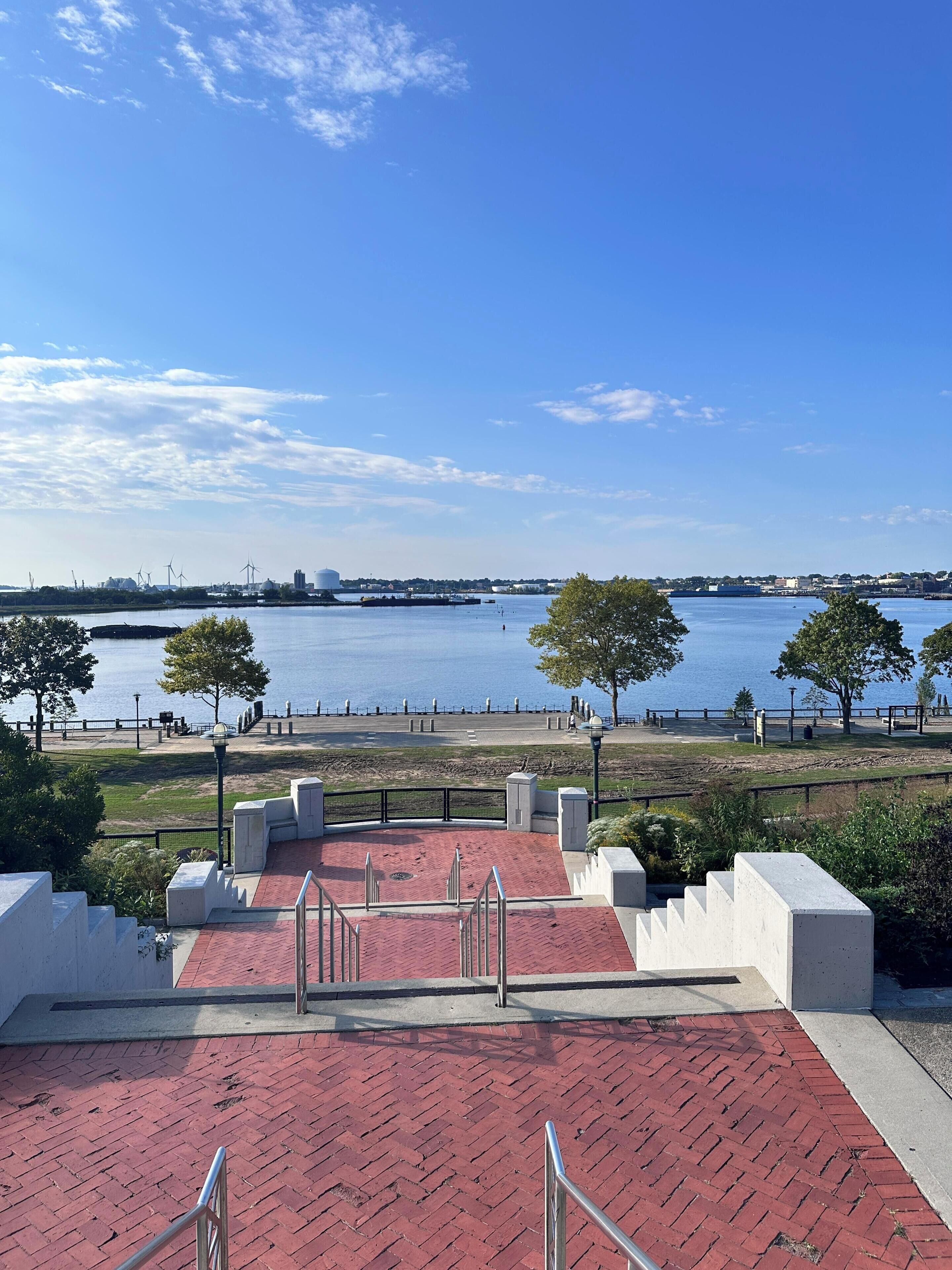 A waterfront park with red brick steps leading to a viewing area, overlooking a calm body of water and distant cityscape.