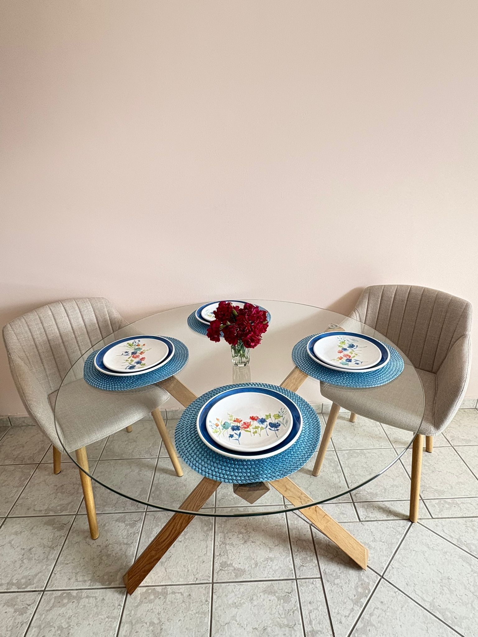 A round glass table with four chairs, each with a blue placemat and a floral plate. A vase with red flowers is on the table.
