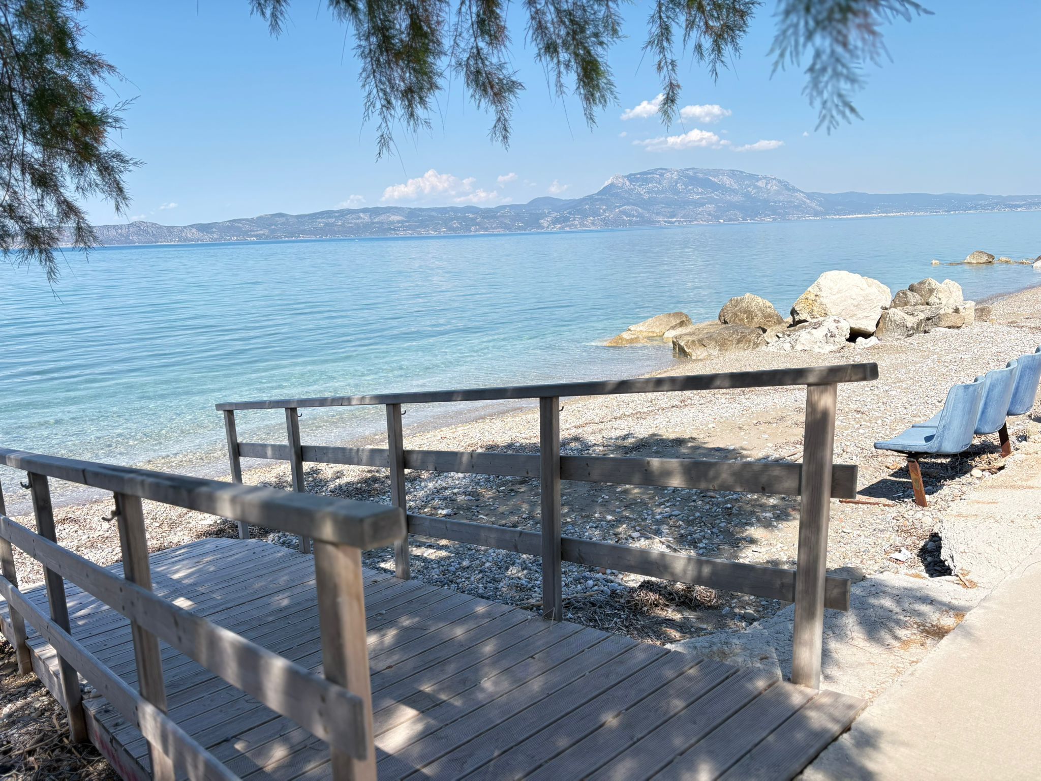 A wooden deck with a railing, pebbles, and a few empty chairs on a beach with clear water and distant mountains.