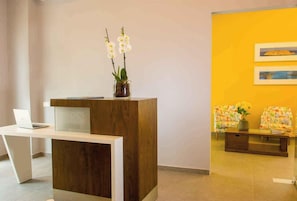 Un mostrador de recepción con una computadora portátil, un área de recepción de madera y una pared amarilla con obras de arte enmarcadas.