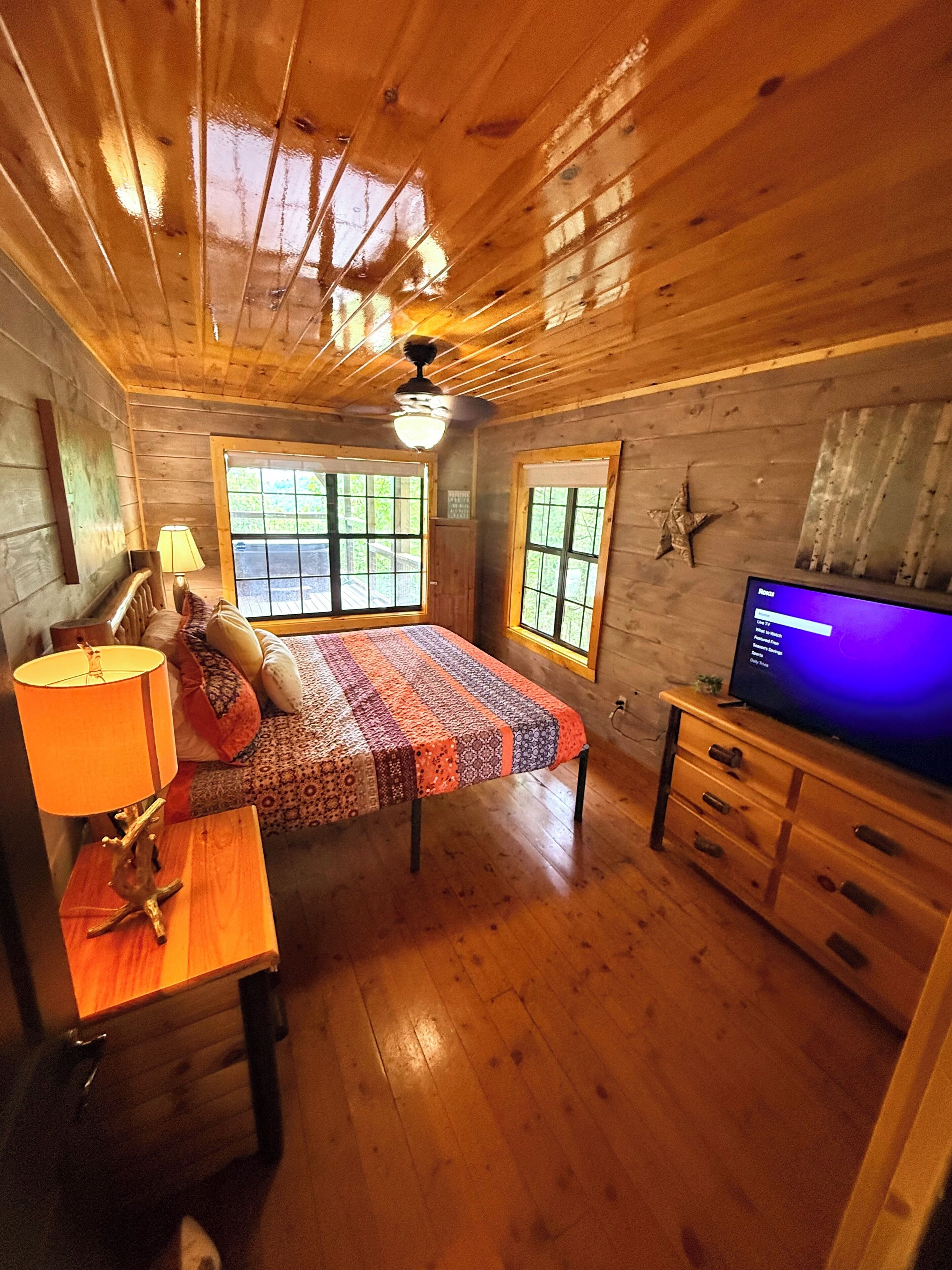 A wooden cabin room with a bed, a desk, a television, and a ceiling fan.