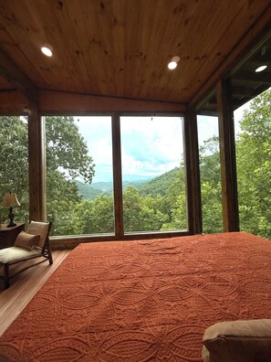 Ein Schlafzimmer mit einem großen Bett, Holzboden und Blick auf eine bewaldete Landschaft durch große Fenster.