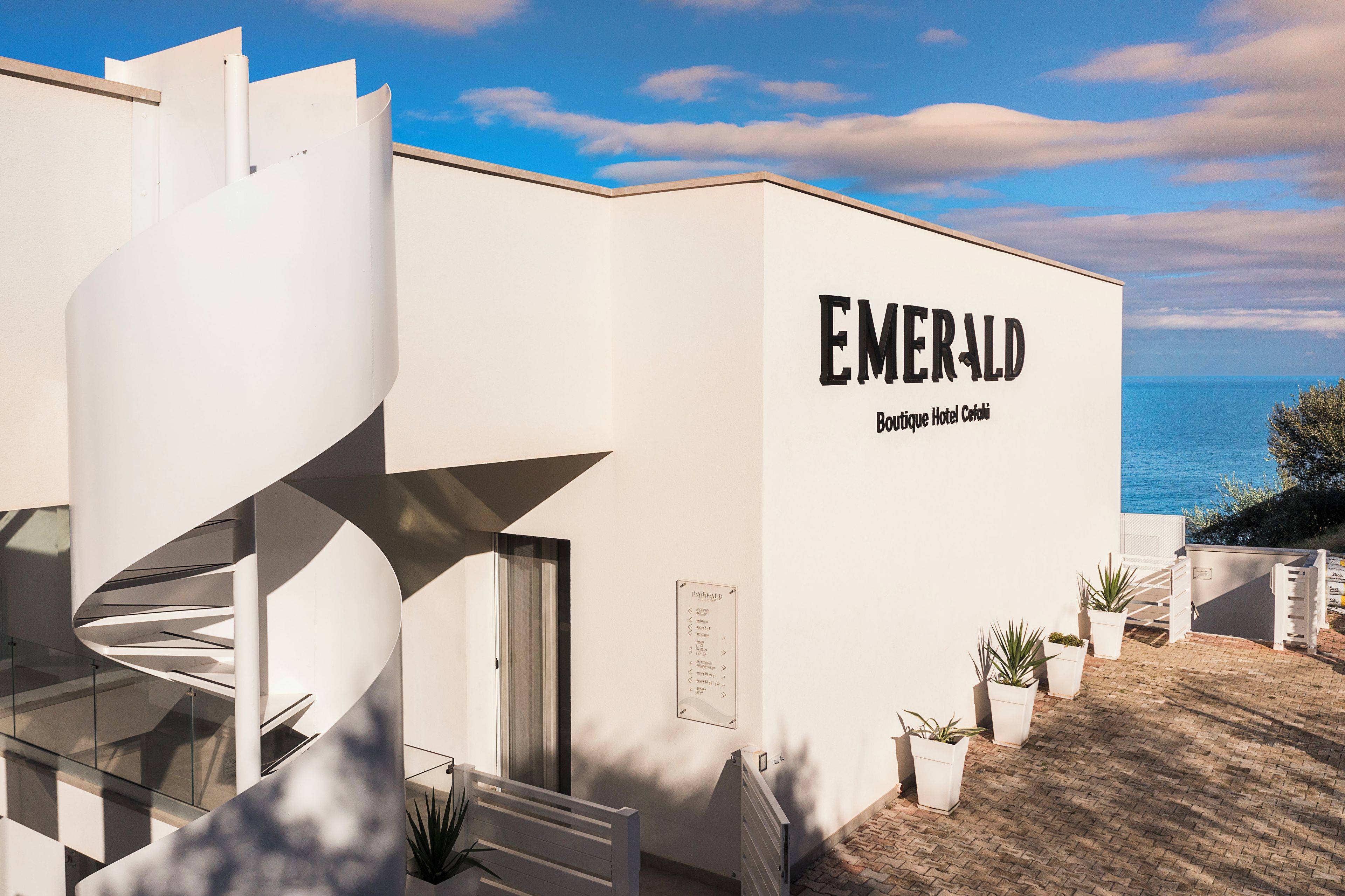 A modern building with a spiral staircase, a sign reading 'EMERALD Boutique Hotel Cartagena,' and a view of the sea.