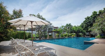 Una piscina con sillones de descanso y sombrillas, rodeada de vegetación y edificios.