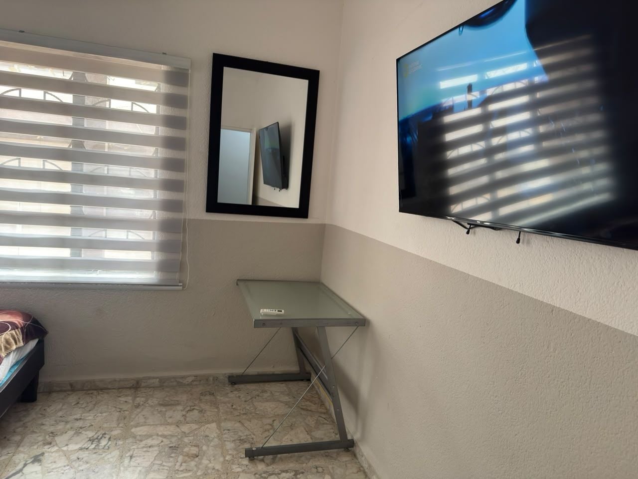 Una habitación pequeña con cama, escritorio, espejo y televisor montado en la pared.