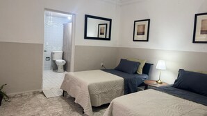 A hotel room with two beds, a bedside table with a lamp, a bathroom with a toilet and shower, and framed pictures on the wall.
