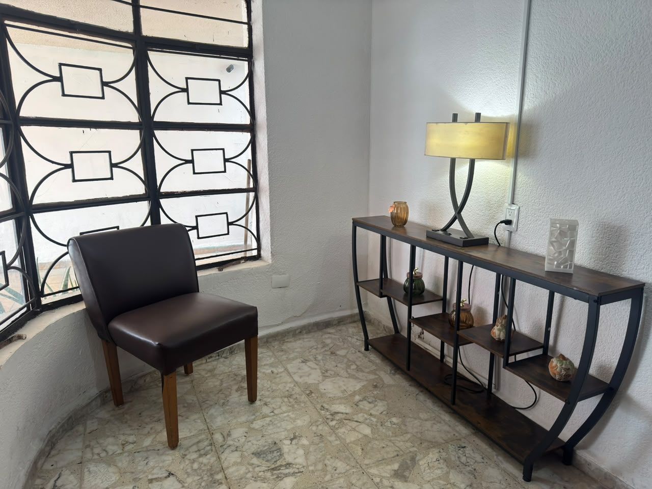 Une pièce comprenant une chaise, une console avec des étagères et une fenêtre ornée d’une grille décorative.