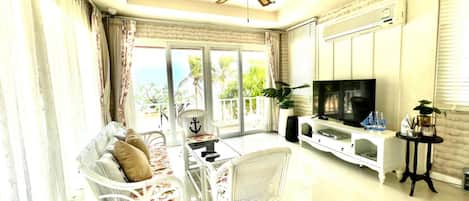 A living room with a TV, white furniture, a ceiling fan, and a view of palm trees.