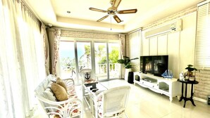 A living room with a TV, white furniture, a ceiling fan, and a view of palm trees. - Rim Chaam By Roomquest (Cha-am)